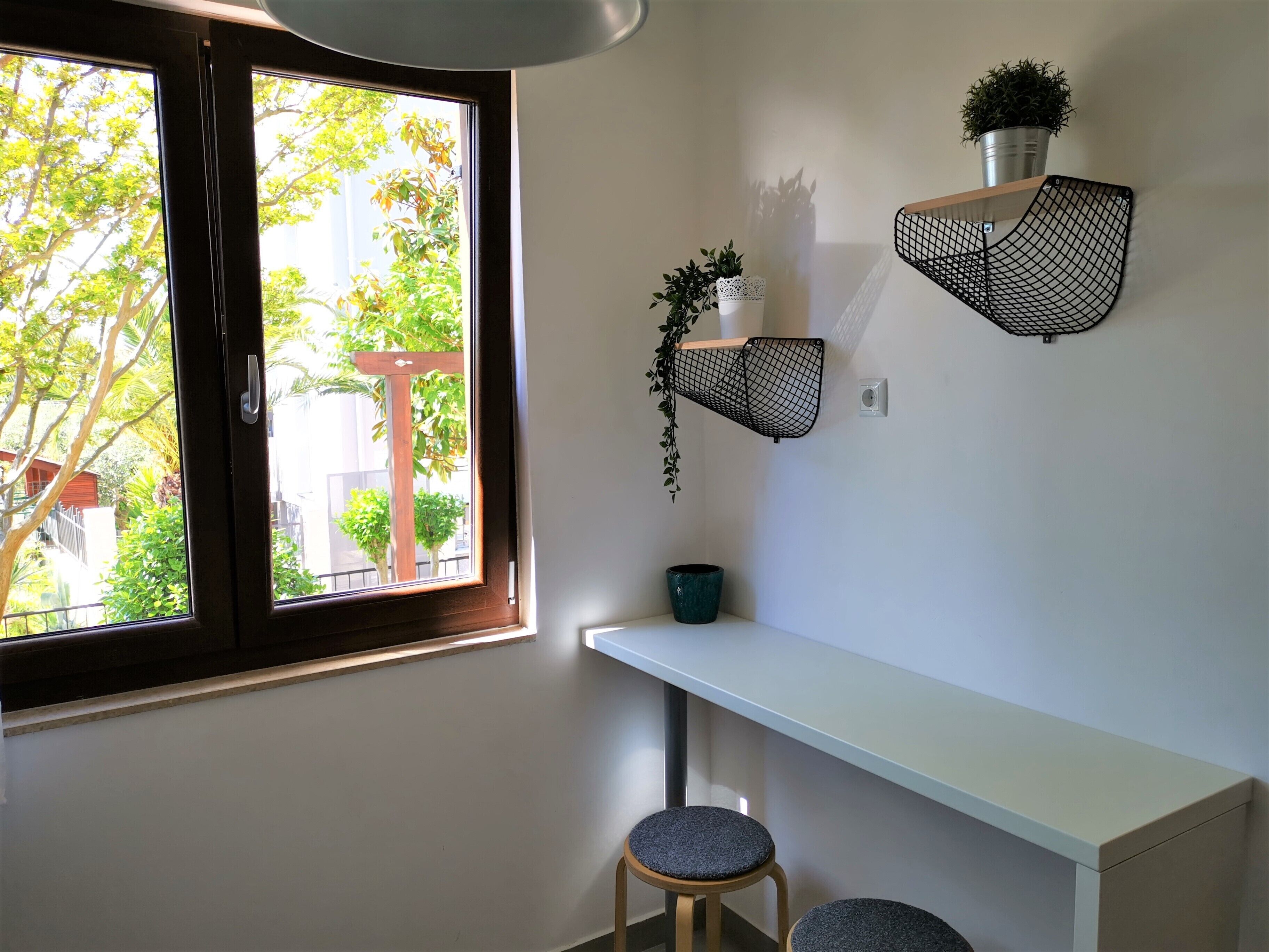 A room with a white desk, two stools, a window with a view of greenery, and two wall-mounted shelves with plants.