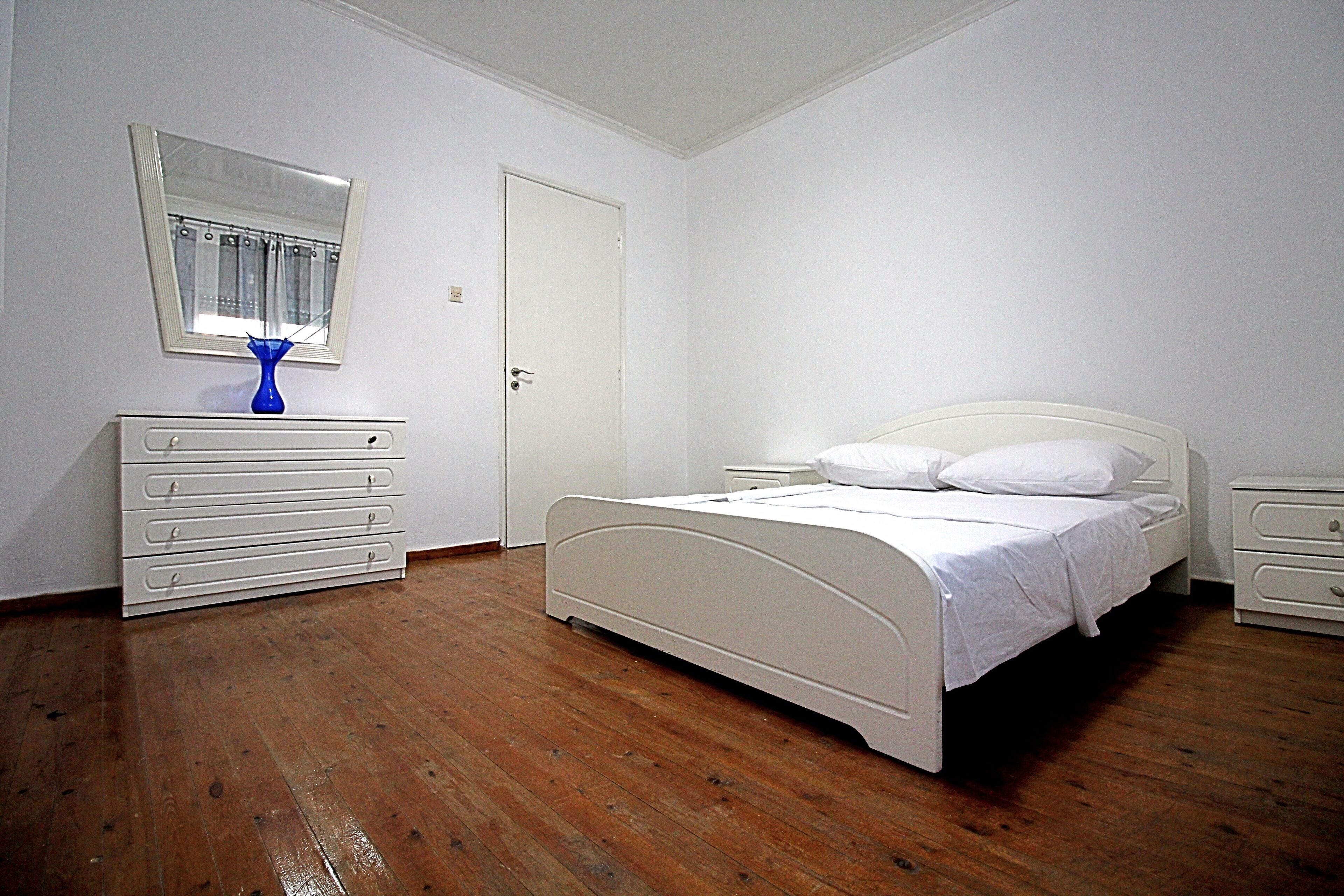 A bedroom with a white bed, a dresser, a nightstand, and a mirror on the wall.