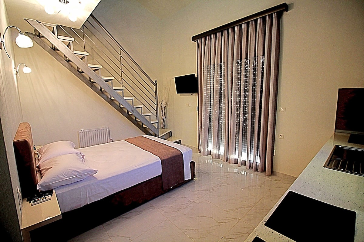 A bedroom with a bed, a nightstand, a desk, a chair, a TV, a window with curtains, a radiator, and a staircase.