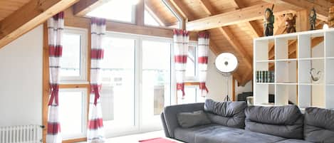 A spacious living room with a grey sectional sofa, wooden ceiling beams, a white shelving unit, and a red table.