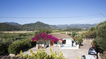 A villa with a tiled roof, surrounded by greenery and mountains in the background.