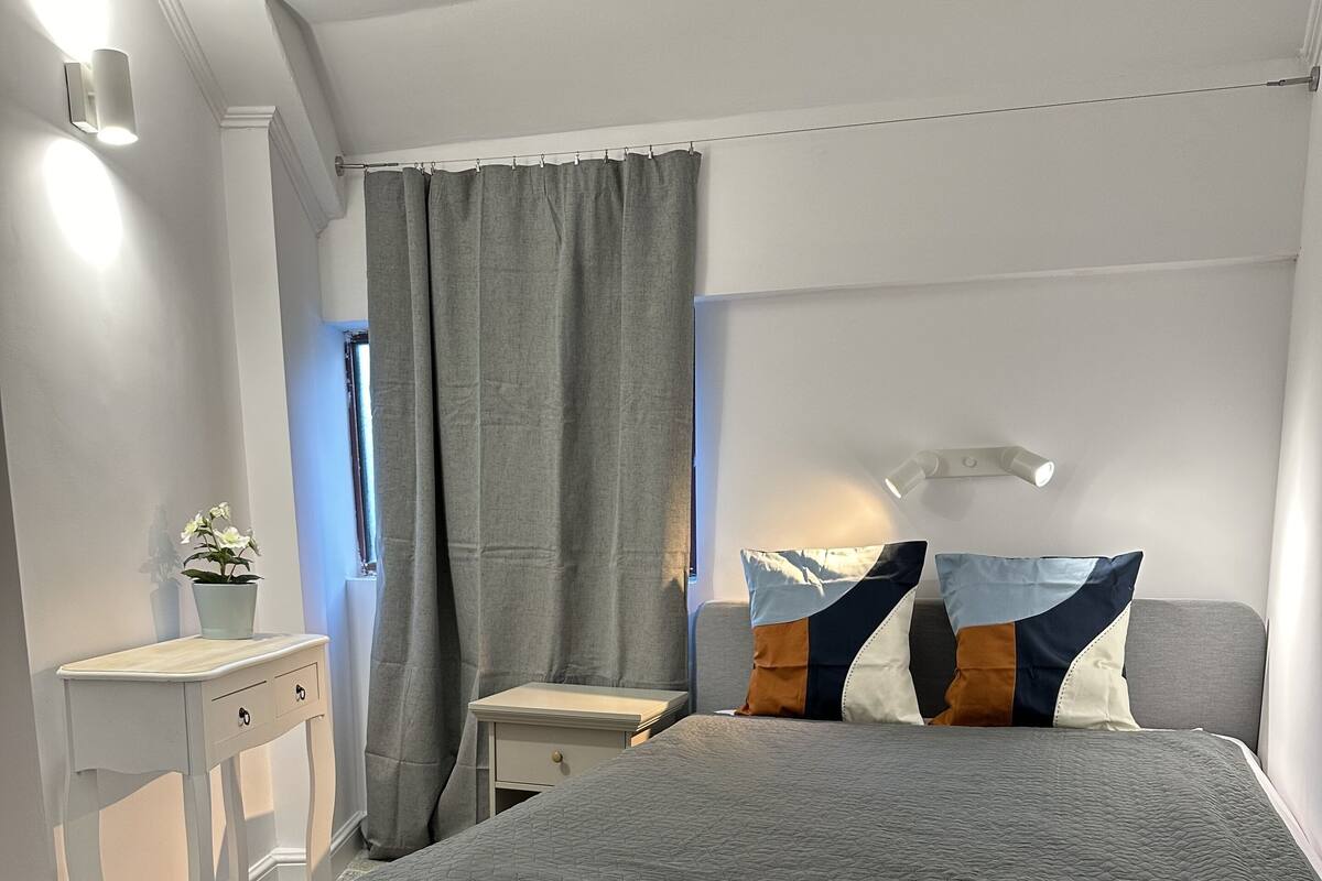 A bedroom with a large bed, a bedside table, a nightstand, a window with curtains, and a wall-mounted light fixture.