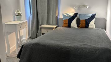 Une chambre à coucher avec un grand lit, une table de chevet, une commode, une fenêtre avec des rideaux et un luminaire fixé au mur.