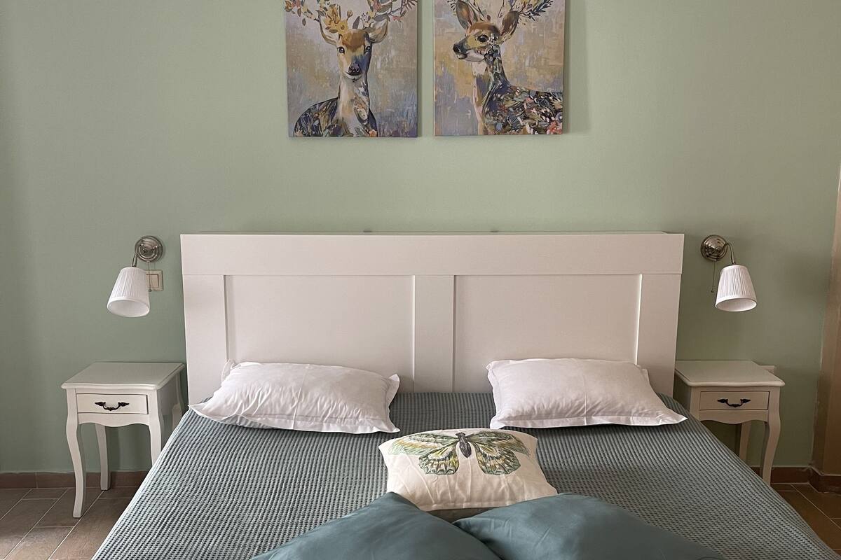 A neatly made bed with a green and white checkered bedspread, two white pillows, and a decorative pillow with a leaf design. There are two wall-mounted paintings of deer, and two bedside lamps on white nightstands.