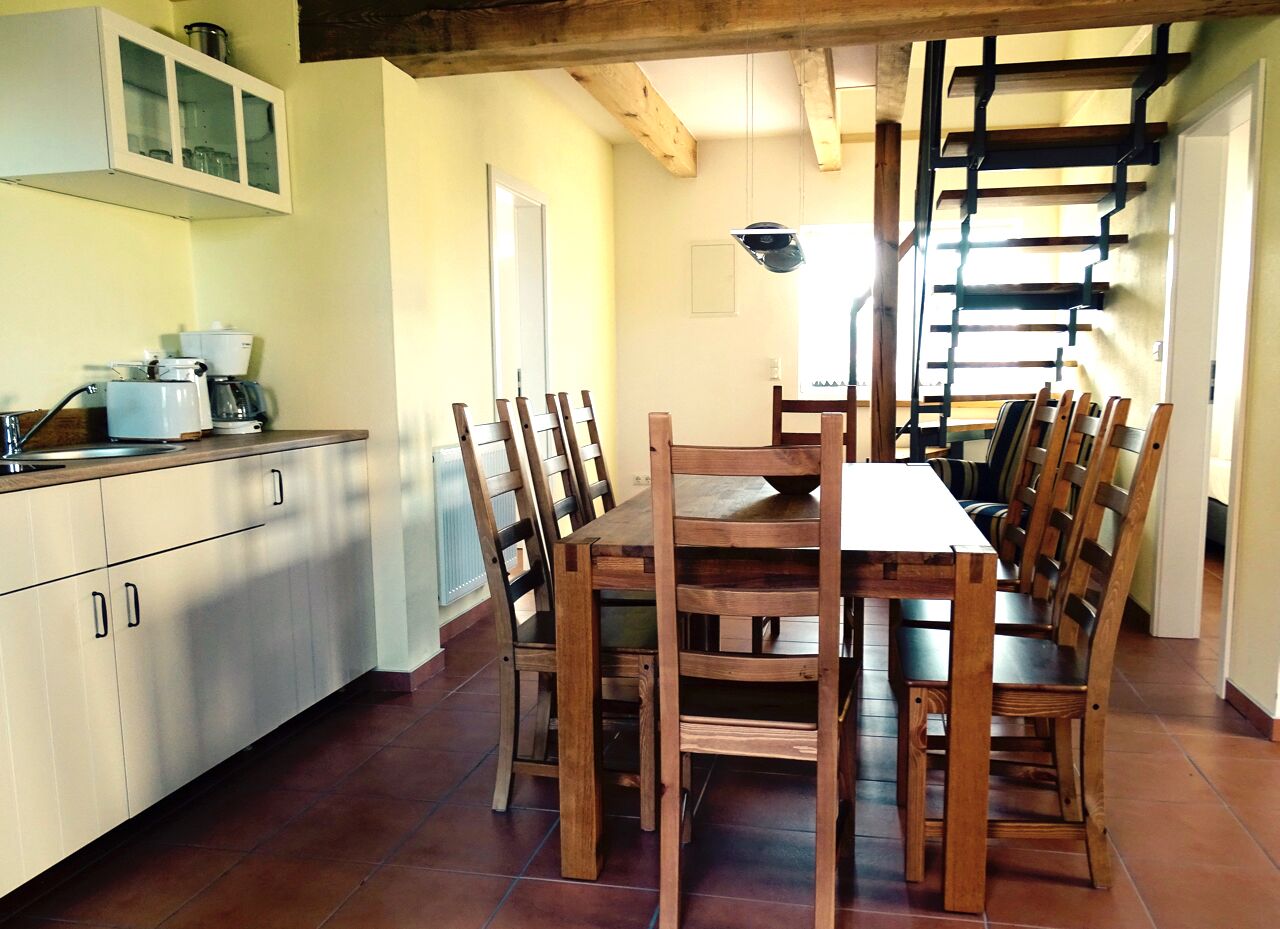 Een eetgedeelte met een houten tafel en stoelen, een keuken met kasten en apparatuur, en een trap naar een ander niveau.