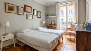 A bedroom with two beds, a desk, a chair, and a window with a view of buildings. - Muted Beauty in Batignolles, by Veeve (Paris)