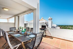 An outdoor dining area with a table set for two, a grill, and a view of the ocean.