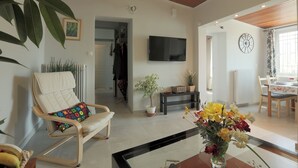 A living room with a TV, a chair with a cushion, a potted plant, a dining area with a table and chairs, and a clock on the wall. - Splendid Views from a Cozy Cottage with a Garden Terrace (Dafni)