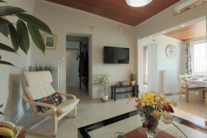 Una sala de estar con televisor, una silla con cojín, una planta en maceta, un comedor con mesa y sillas, y un reloj en la pared.