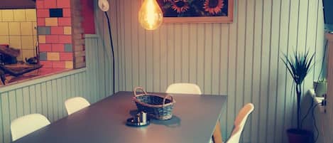 Un comedor con una mesa rectangular, seis sillas blancas, una bombilla colgante, una cesta y un cuadro en la pared.