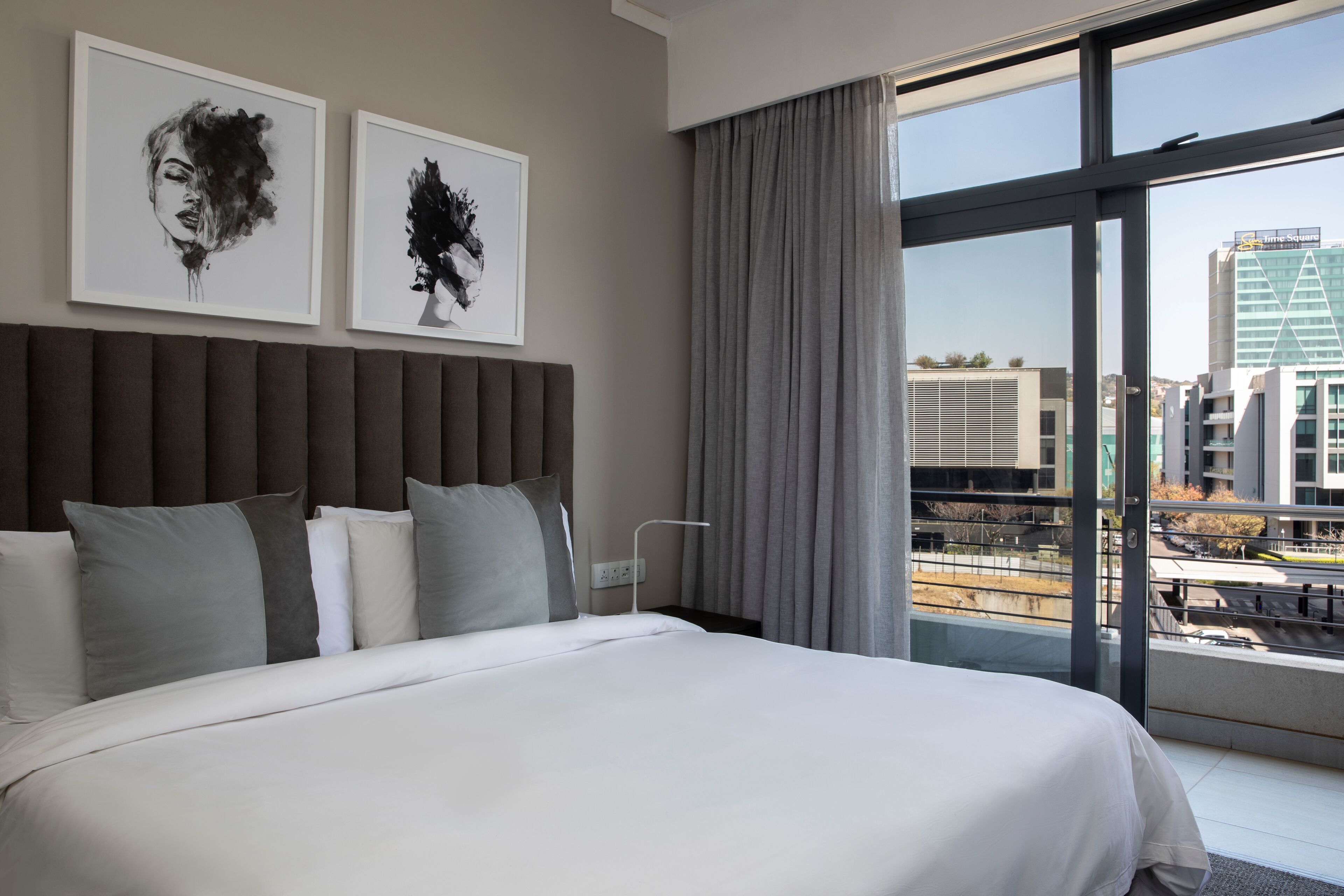 Una habitación de hotel moderna con una cama grande, ropa de cama gris y blanca, dos cuadros en la pared y vistas de la ciudad a través de amplias ventanas.