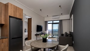 A modern kitchen with a black refrigerator, wooden cabinets, a dining area with a marble table and chairs, a flat-screen TV, and a large window with a city view. - The Capital Trilogy (Pretoria)