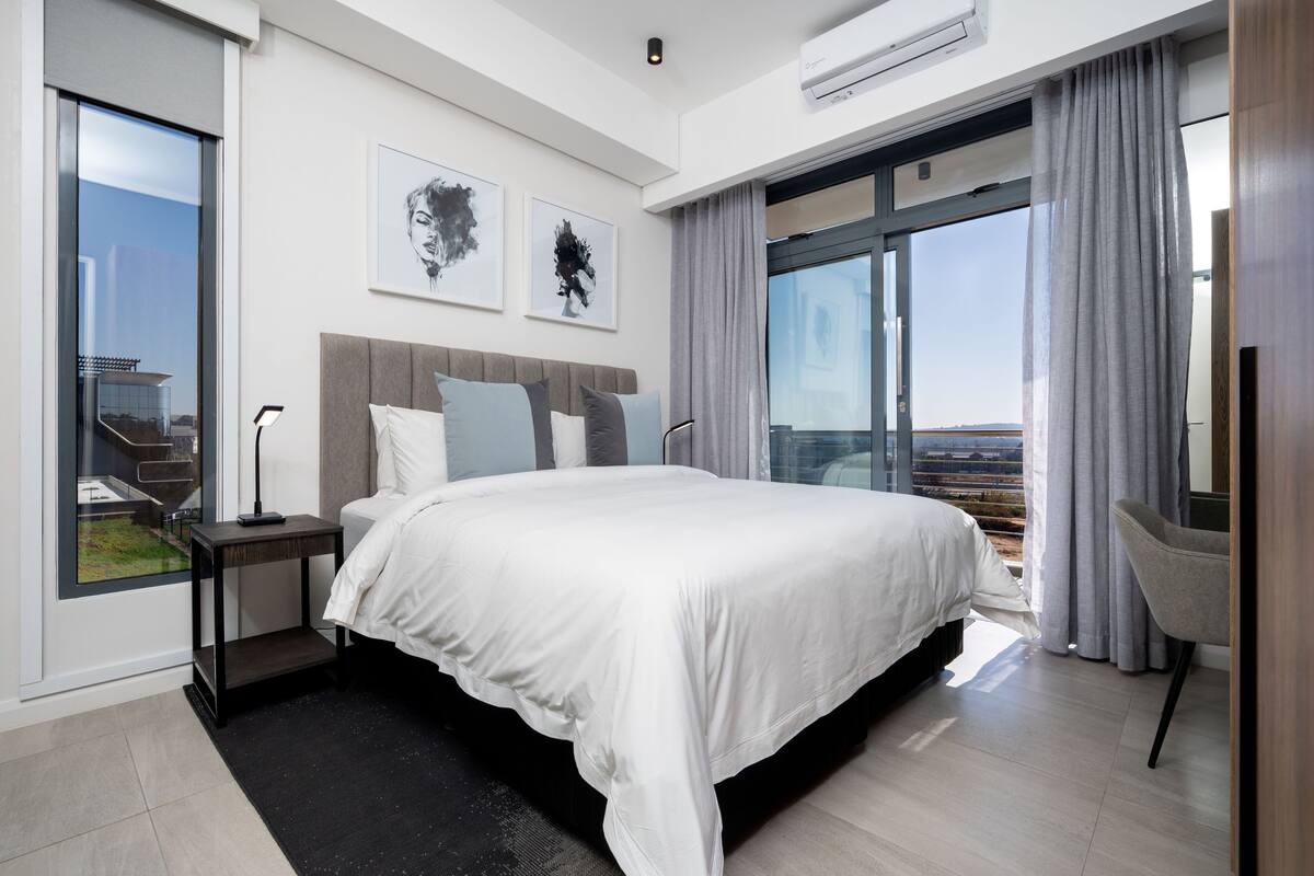 A modern hotel room with a large bed, a nightstand, a chair, and a view of the city through large windows.
