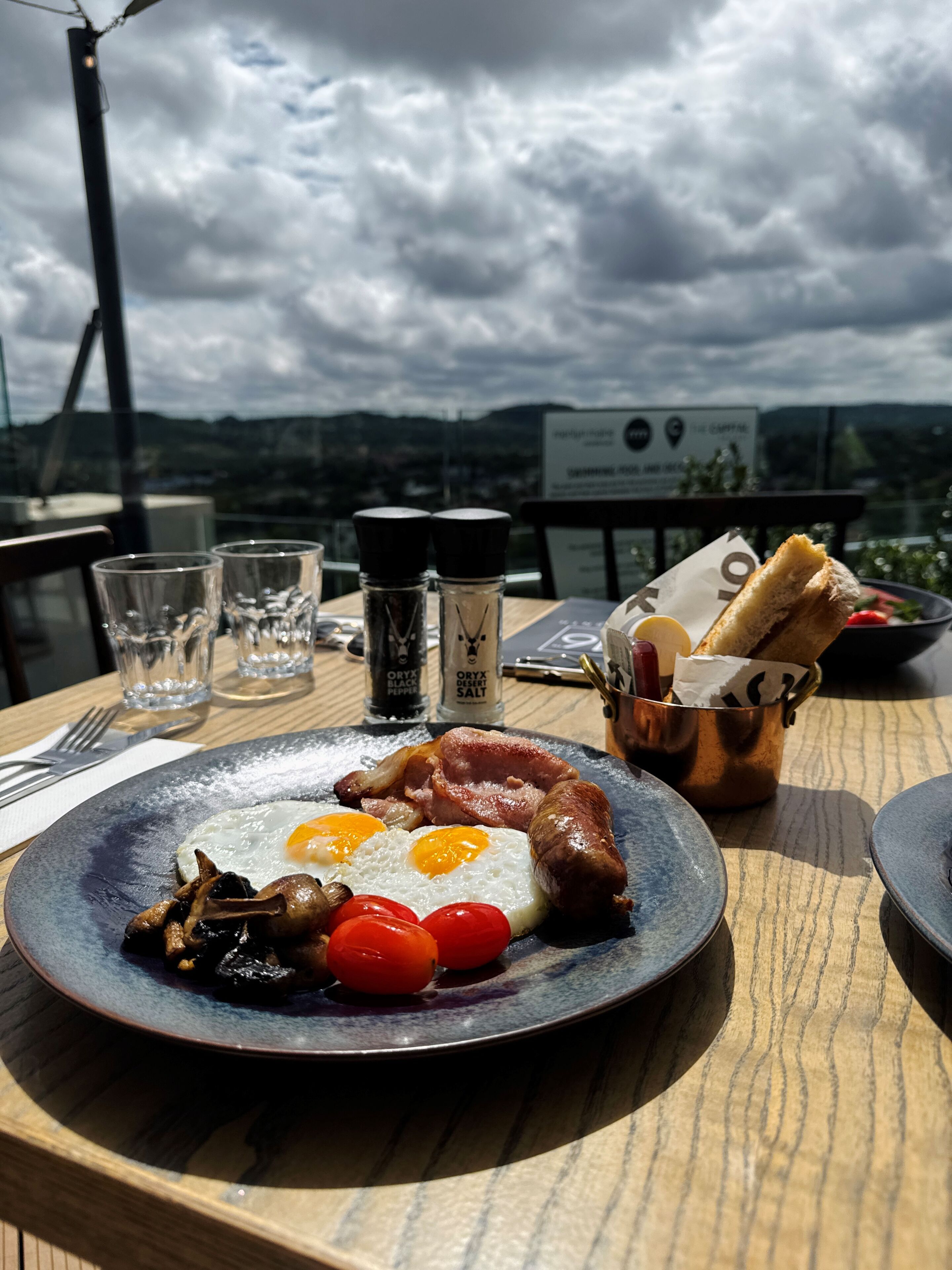 A breakfast plate with eggs, bacon, mushrooms, and tomatoes on a blue plate, with salt and pepper shakers and a bread basket on a wooden table.