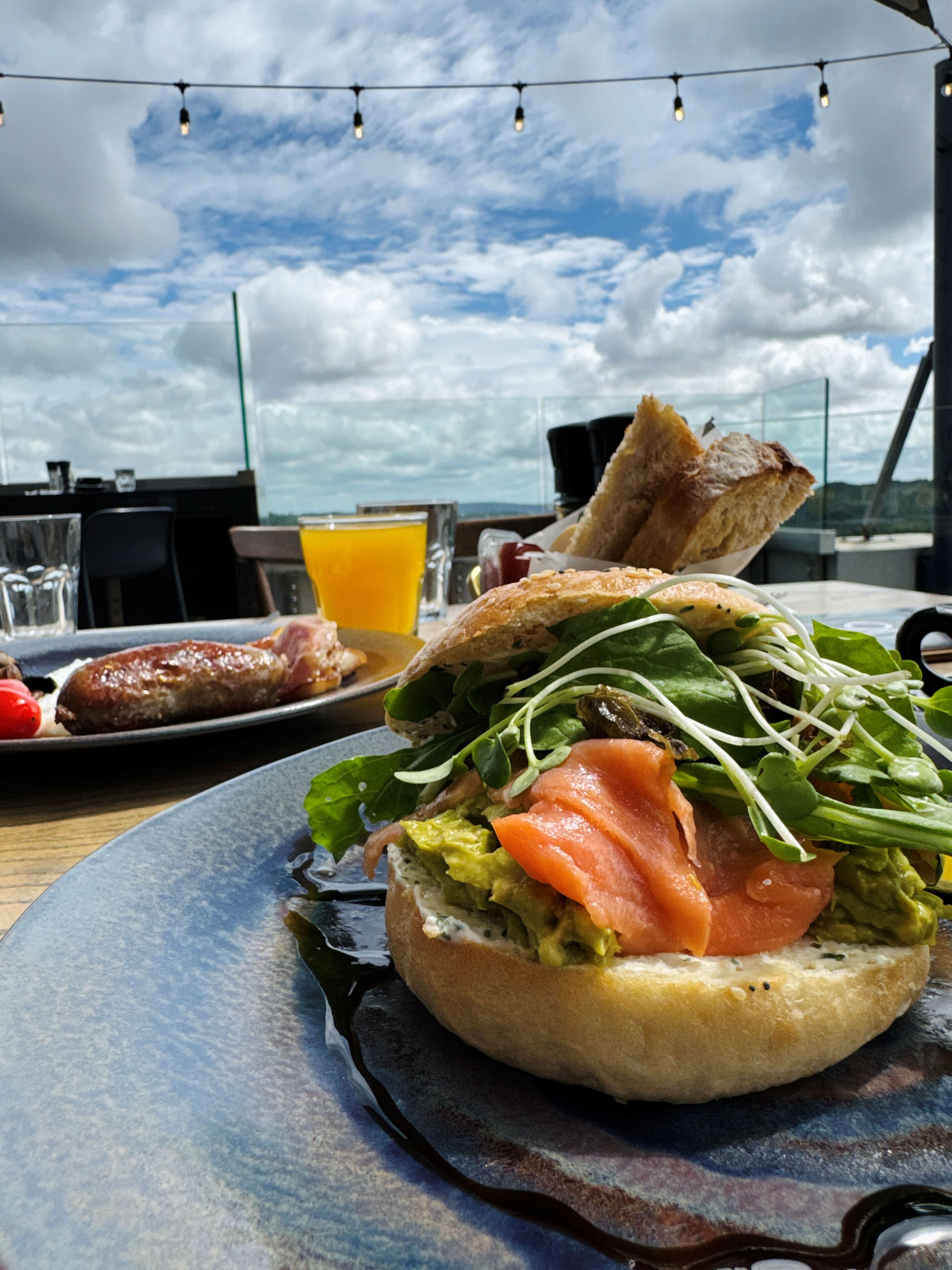 Un sándwich de aguacate y salmón ahumado sobre un plato, un vaso de jugo de naranja y una rebanada de pan en una mesa con vista al cielo.