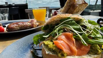 Un sándwich de aguacate y salmón ahumado sobre un plato, un vaso de jugo de naranja y una rebanada de pan en una mesa con vista al cielo.