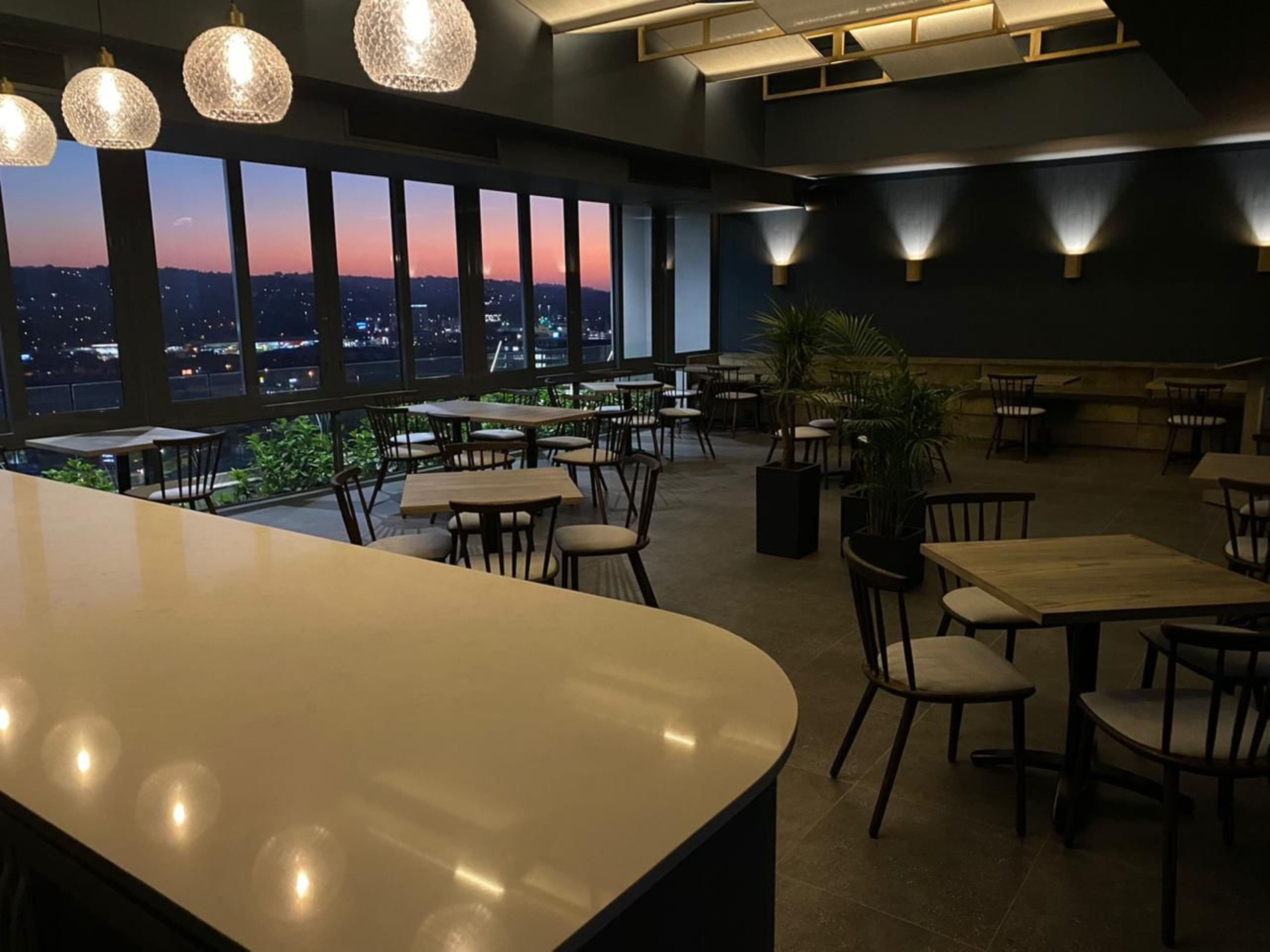 Un restaurant doté d’un bar, de nombreuses tables, des chaises et de grandes fenêtres offrant une vue sur le paysage urbain au crépuscule.