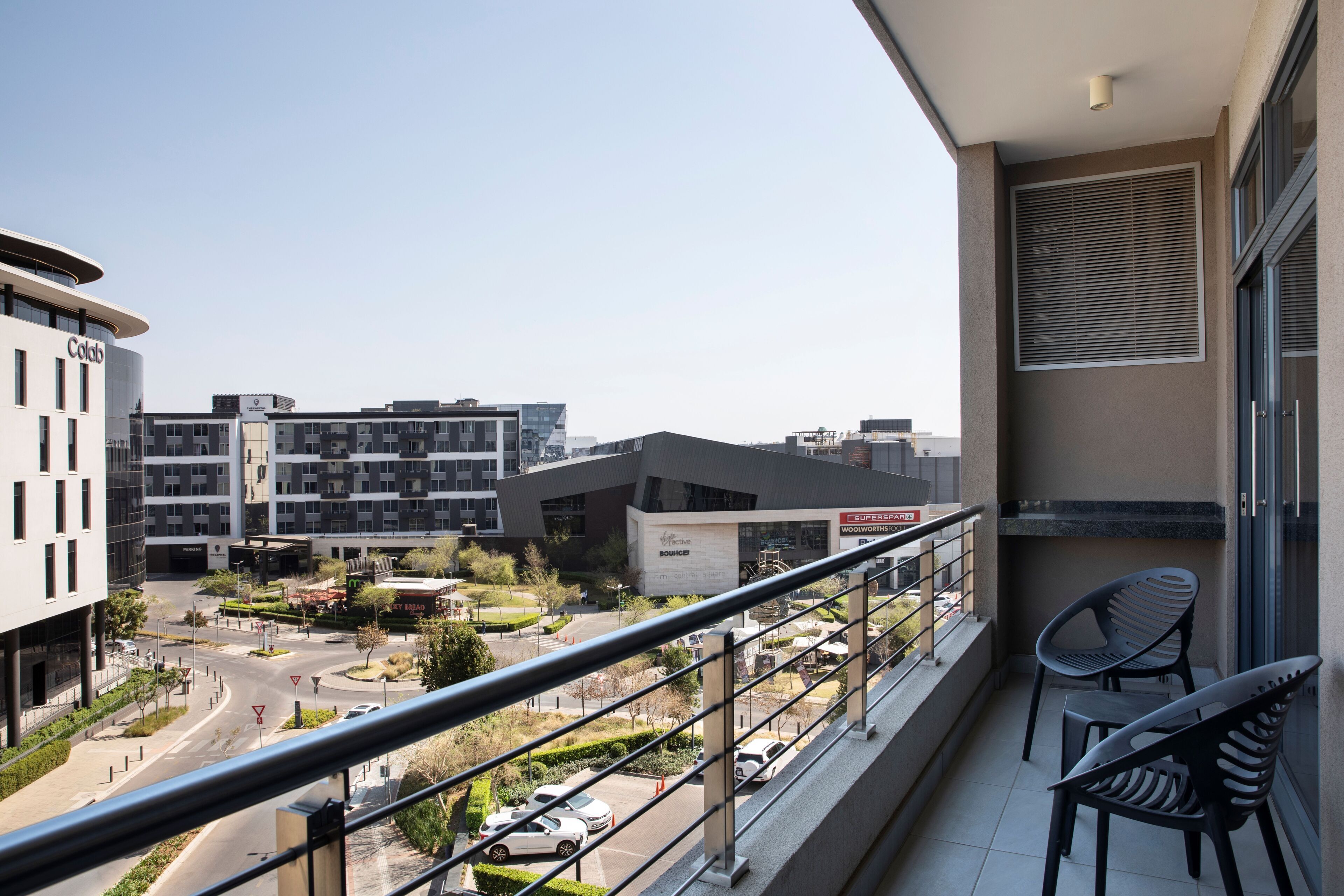 A balcony with two chairs, a view of a cityscape with modern buildings, and a road with cars.