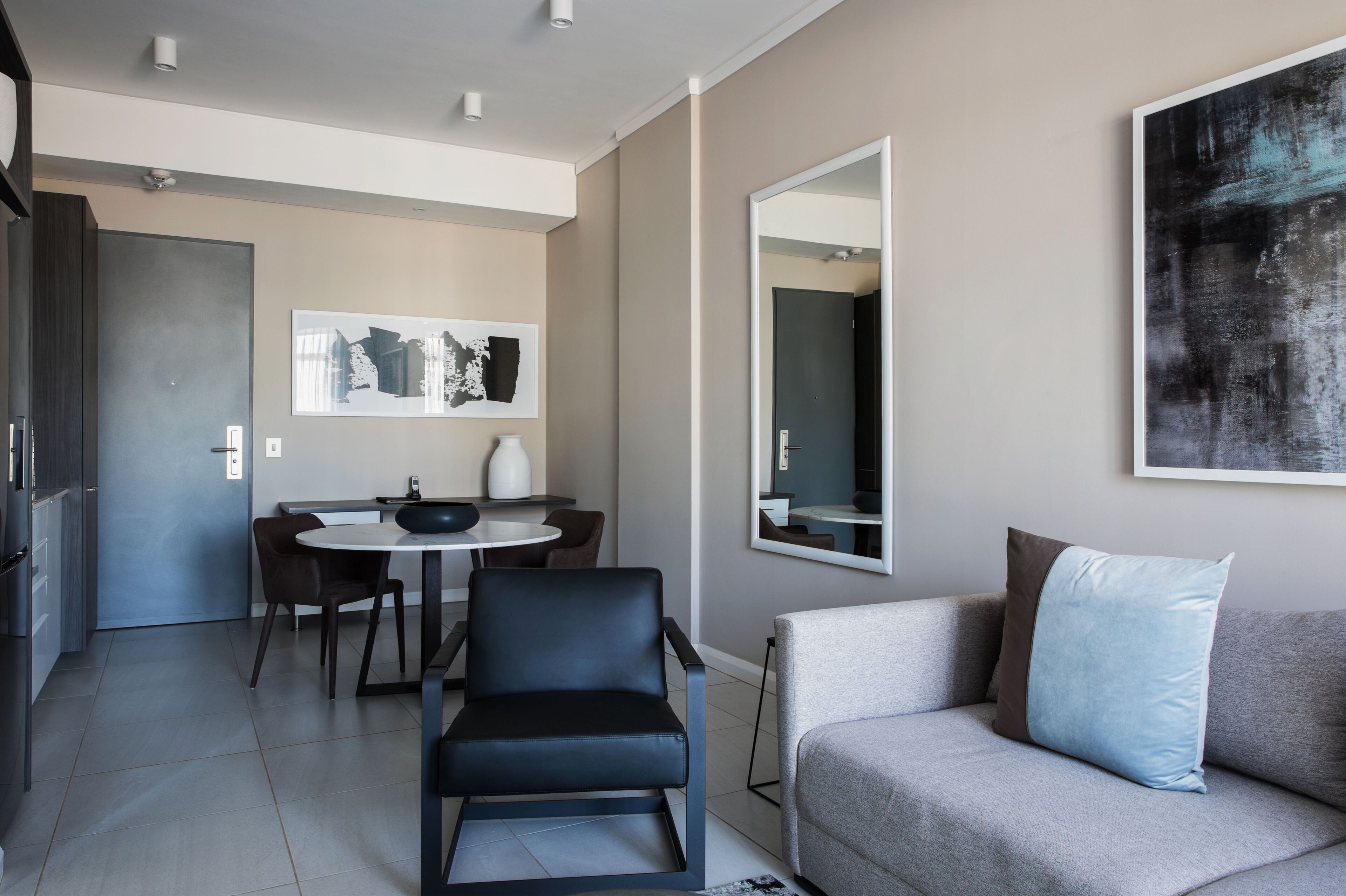 A modern living room with a grey sofa, a black chair, a round dining table, and a large abstract painting on the wall.