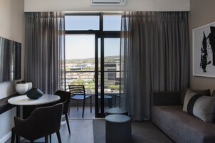 A modern hotel room with a balcony, a sofa, a dining table with chairs, and a large window with curtains.