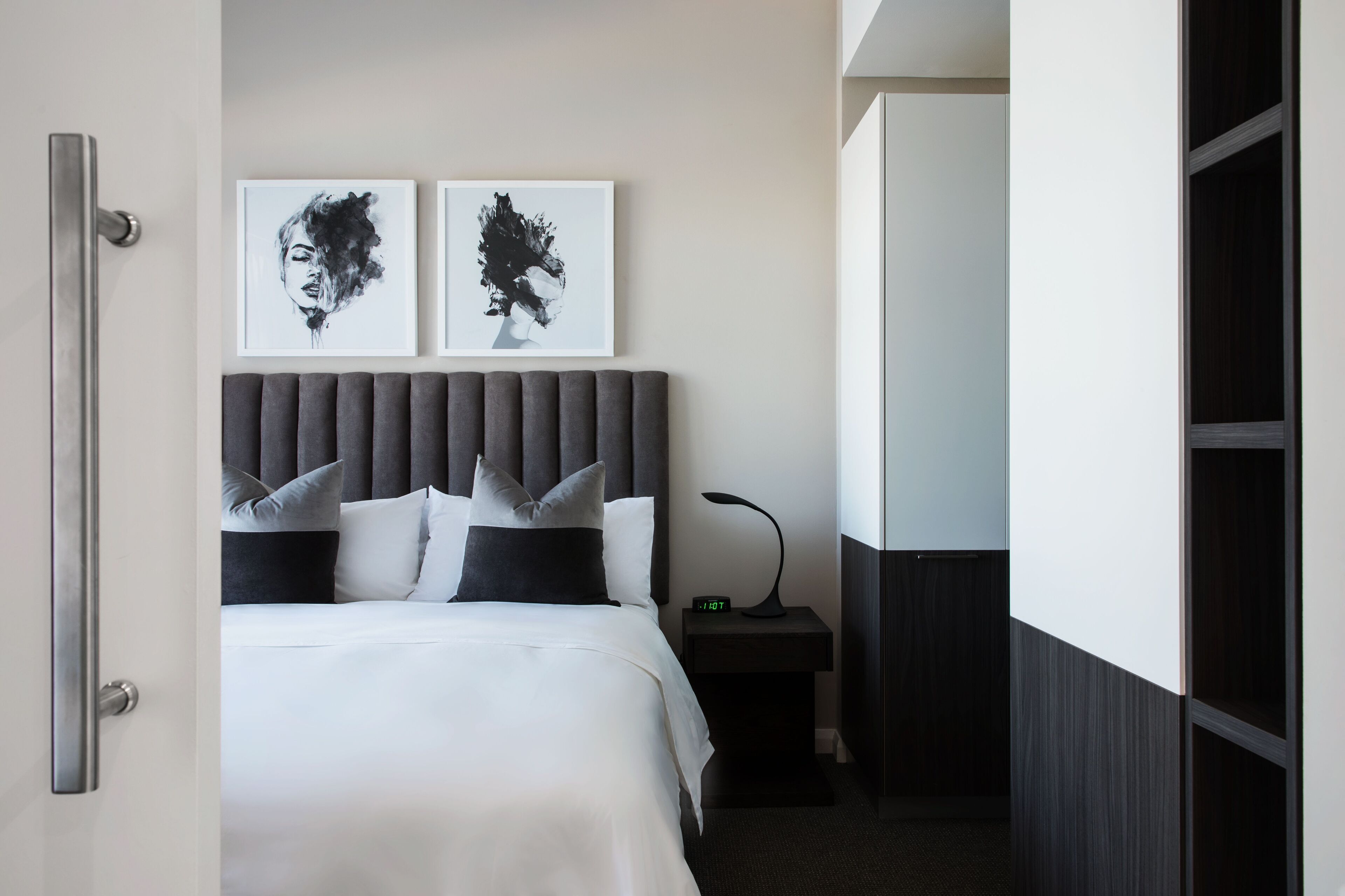 A hotel room with a bed, two framed artworks on the wall, a nightstand with a lamp, and a wardrobe.
