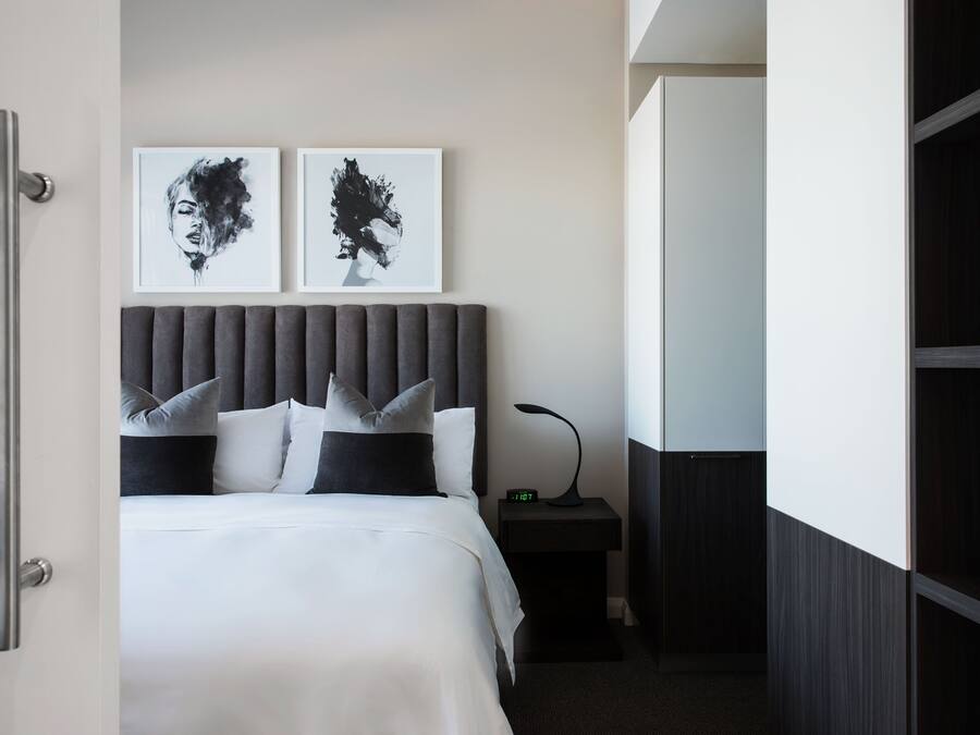 A hotel room with a bed, two framed artworks on the wall, a nightstand with a lamp, and a wardrobe.