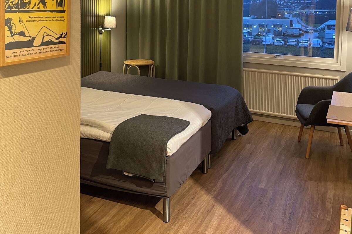 Quarto de hotel com duas camas, uma janela com vista para a cidade, um cartaz na parede e uma pequena mesa com uma cadeira.