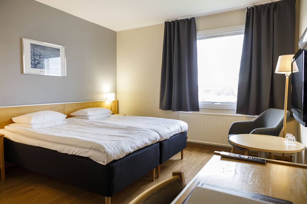 Quarto de hotel com cama, escrivaninha, cadeira, televisão e janela com cortinas.