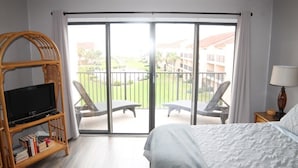 Een hotelkamer met een bed, een televisie op een standaard, een balkon met stoelen en uitzicht op gebouwen en groen.