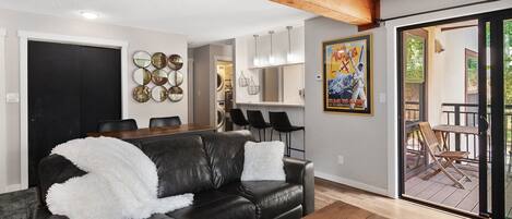 A living room with a black leather sofa, a wooden coffee table, and a dining area with a black table and chairs. There is a kitchen area with a refrigerator and a poster on the wall.