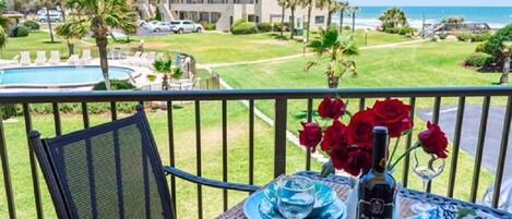 A balcony with a table set for two, overlooking a pool and a building with a view of the ocean.