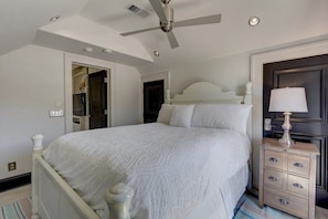 Une chambre à coucher comprenant un lit, une table de chevet, une lampe, un ventilateur de plafond et une porte donnant sur une autre pièce.