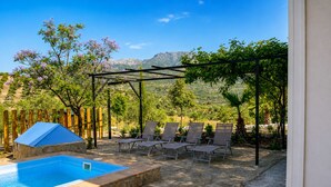 Ein Pool mit Liegestühlen und einer Pergola, umgeben von viel Grün und Bergen im Hintergrund.