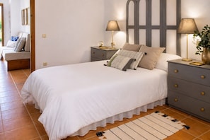 A bedroom with a large bed, two bedside tables with lamps, a nightstand, a wall-mounted clock, and a door leading to another room.
