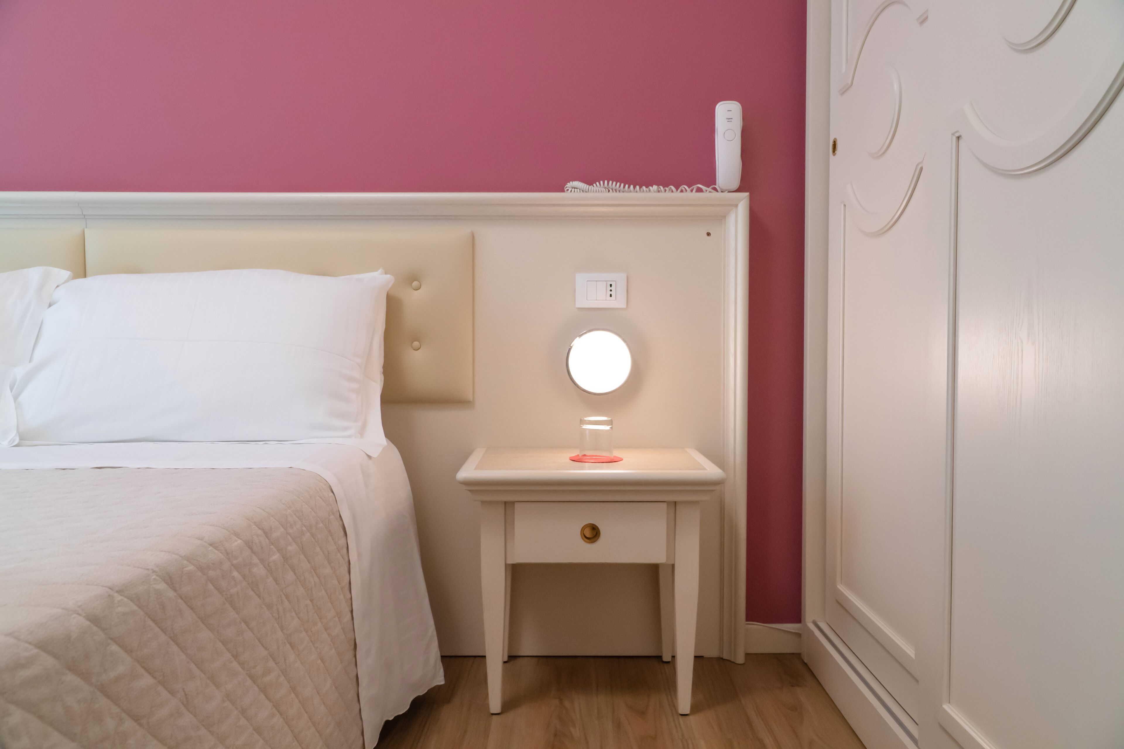 A neatly made bed with a white comforter and pillows, a bedside table with a round mirror, a small drawer, and a wall socket.