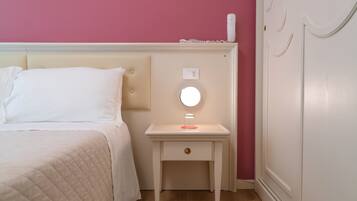 A neatly made bed with a white comforter and pillows, a bedside table with a round mirror, a small drawer, and a wall socket.