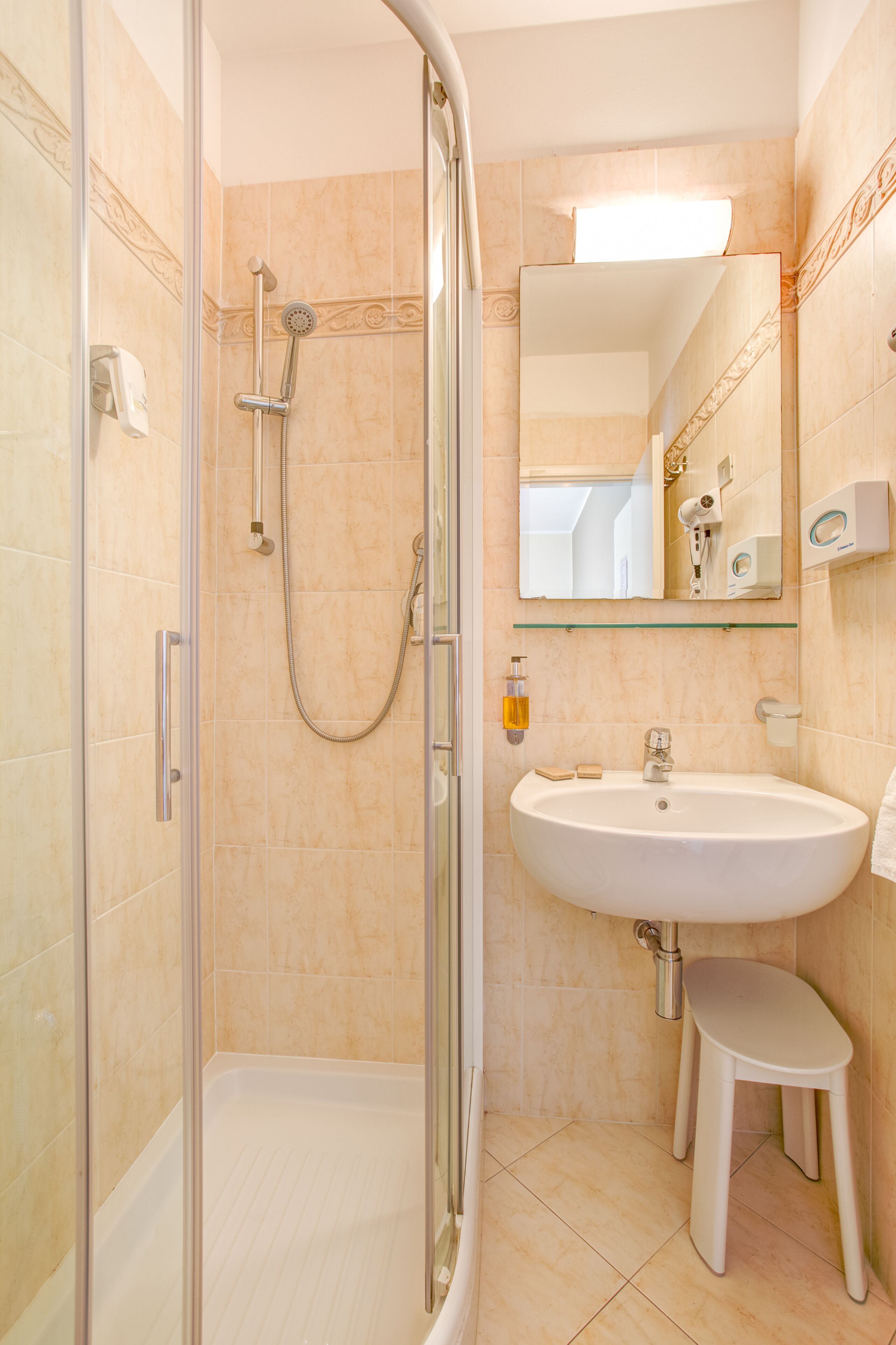 Un baño con ducha de vidrio, lavamanos, espejo y un taburete pequeño.