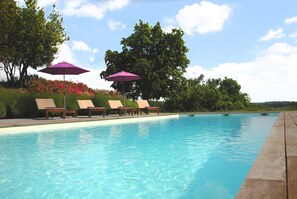 Una piscina con sillas de descanso y sombrillas, rodeada de follaje y árboles.