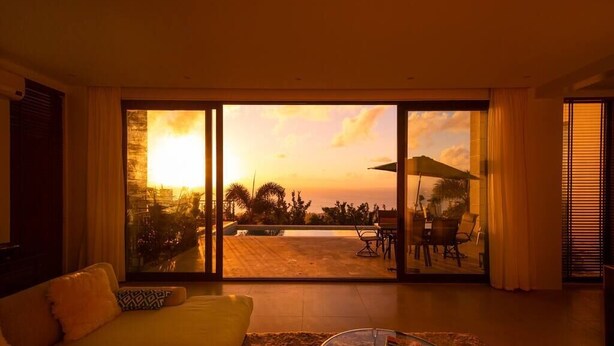 A living room with a sofa, a coffee table, and a view of a patio with outdoor furniture and a sunset. - Navy Hill Resort (Saipan)