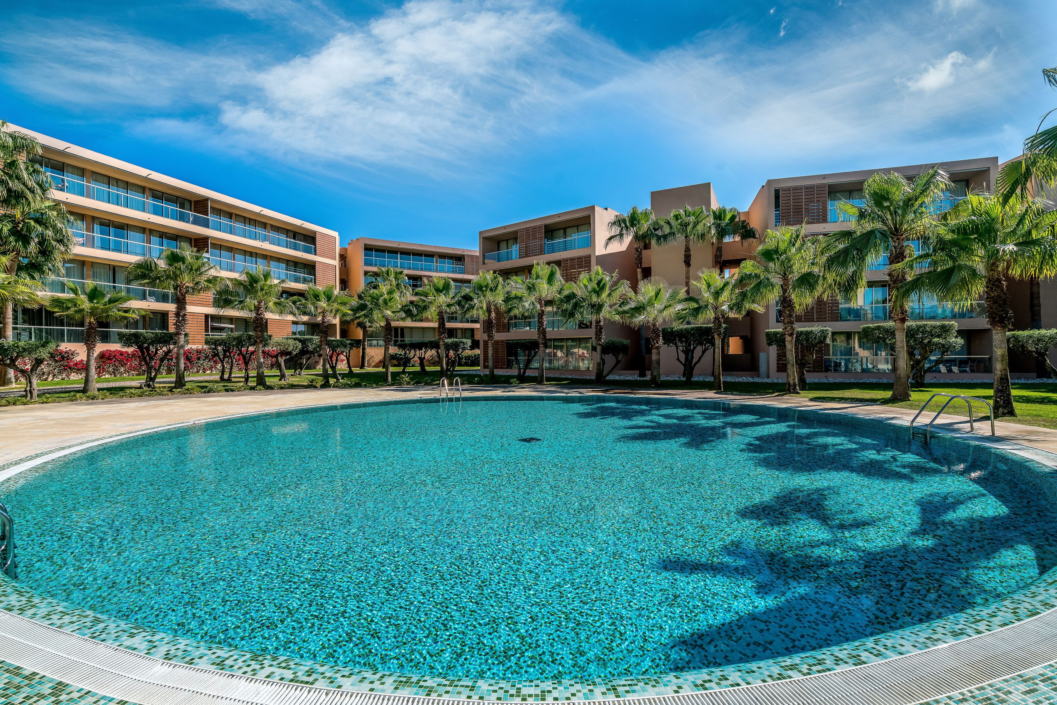 A large, clear swimming pool surrounded by modern buildings with multiple floors and balconies, palm trees, and a clear blue sky.