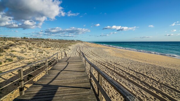 Ein hölzerner Steg führt zu einem Sandstrand, im Hintergrund strahlend blauer Himmel und das Meer.