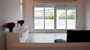 A modern room with a wooden ceiling, a white console table with red flowers, a black sofa, and large windows with a view of buildings and greenery. - Hostal Central (Fuente Alamo)