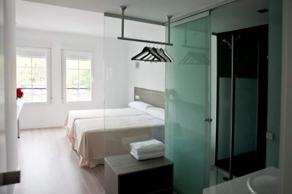 A modern hotel room with two beds, a glass shower enclosure, and a view of the outdoors.