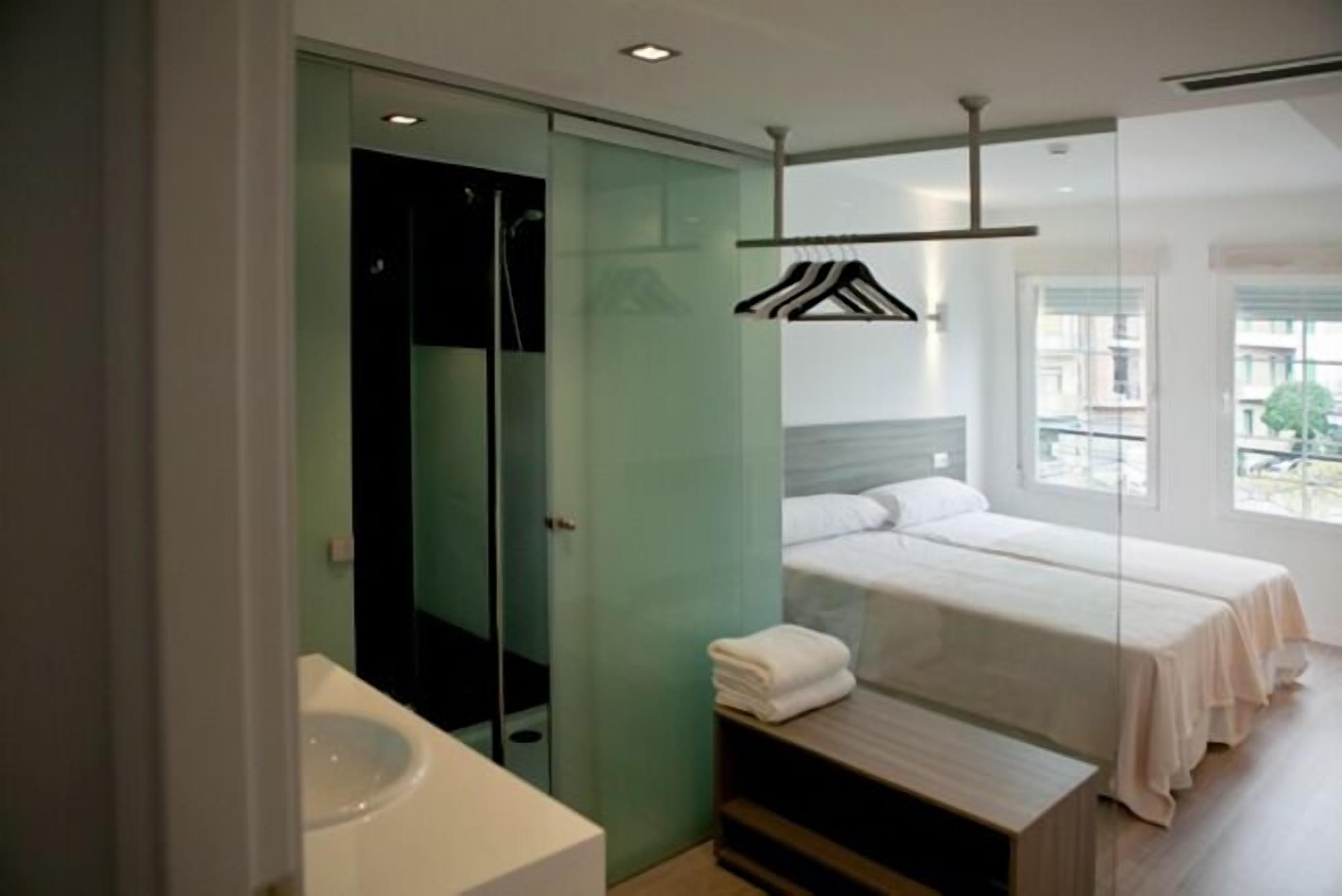 A modern hotel room with a glass shower enclosure, a bed with white linens, and a view of the city through large windows.