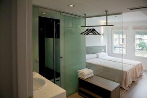 A modern hotel room with a glass shower enclosure, a bed with white linens, and a view of the city through large windows.
