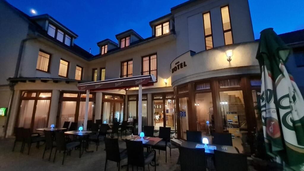 Ein Außenbereich mit Tischen und Stühlen, ein überdachter Bereich und im Hintergrund ein Gebäude mit der Aufschrift 'HOTEL'.