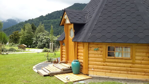 Une cabane en rondins de bois, dotée d’un toit hexagonal, d’un porche en bois et d’un grand vase vert.
