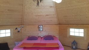 Um quarto de cabana de madeira acolhedor, com uma cama, duas janelas, um quadro e um candeeiro.