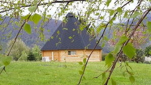 Uma cabana de tronco com telhado cónico, rodeada por um campo gramado e montanhas ao fundo.