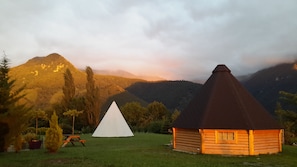 Uma cabana com telhado cónico e uma tenda branca numa área gramada, com montanhas ao fundo.